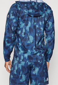 Blå och teal-mönstrad vindjacka med huva, elastiska ärmslut och nederkant. Tillverkad av lätt, vattenavvisande material.