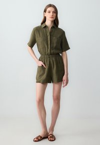 Olivengrüner Leinen-Short-Jumpsuit mit kurzen Ärmeln, Knopfleiste vorne, elastischem Bund und zwei Brusttaschen. Kombiniert mit braunen flachen Sandalen.