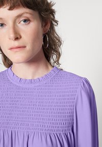 Top smock de lavanda com decote franzido e mangas longas, apresentando um corpete texturizado e tecido suave. Brincos de argola prateados são visíveis.