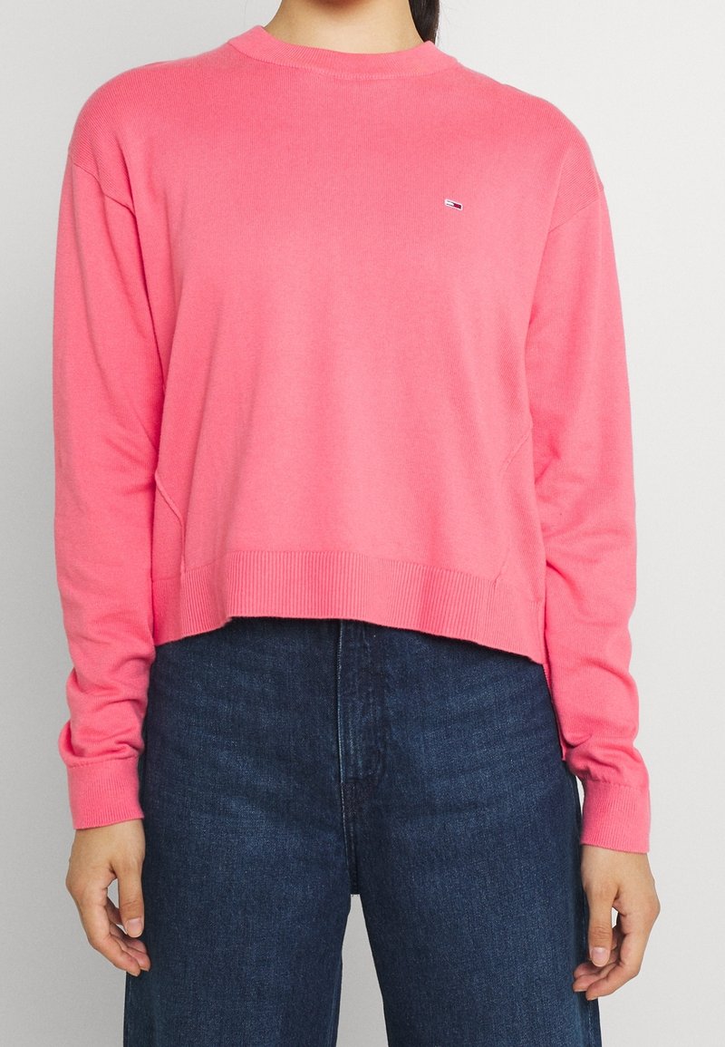 Femme portant un pull rose vif à manches longues avec poignets et ourlet côtelés, associé à un jean taille haute bleu foncé.