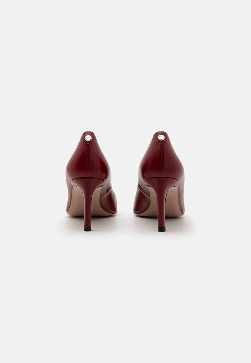 Talons hauts en cuir verni rouge avec un bout pointu, un talon slim aiguille, et un petit accent argenté à l'arrière du talon. Texture lisse.