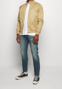 Vuxen som bär en beige jacka, vit t-shirt, slitna blå jeans, vita strumpor och svarta låga sneakers stående mot en enhetlig bakgrund.