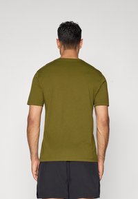 Mann mit dem Rücken zu sehen, trägt ein olivgrünes Kurzarm-T-Shirt und schwarze Shorts vor einem einfarbigen hellen Hintergrund.