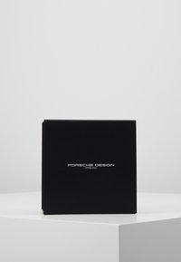 Boîte noire avec finition mate, présentant un logo argenté "Porsche Design" à l'avant. Forme carrée, texture lisse, design minimaliste.