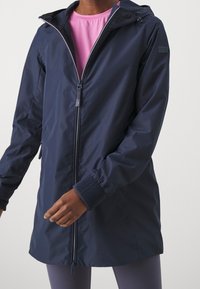 Marinblå vattentät jacka med dragkedja fram, justerbar huva och ribbade ärmslut. Den släta ytan och den något längre designen förbättrar skyddet.