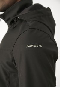 Veste imperméable noire avec capuche et logo "ICEPEAK" sur la partie supérieure de la manche, portée par une personne tournée vers la gauche.