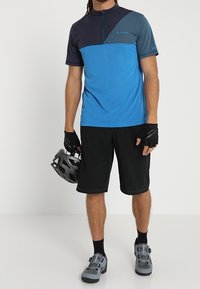 Modré a černé cyklistické tričko s krátkým rukávem a polozipem, v kombinaci s černými šortkami a šedými cyklistickými botami, doplněné o rukavice a helmu.