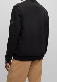 Svart sweatshirt med rund hals, ribbad manschett och nederkant; har en märkespatch på vänster arm; slät textur.