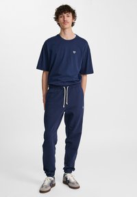 Maglietta in cotone blu navy con un piccolo logo bianco, abbinata a pantaloni sportivi coordinati con polsini elasticizzati e vita con cordoncino. Sneakers neutre.