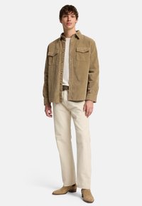 Camicia in corduroy color tan con due tasche sul petto, abbinata a pantaloni off-white e stivali in suede marrone chiaro. Il modello è in piedi su uno sfondo neutro.