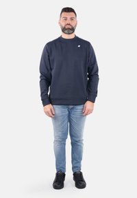Felpa blu navy con colletto girocollo e piccolo logo, abbinata a jeans azzurri chiari e sneakers nere. Presenta maniche lunghe e una vestibilità comoda.