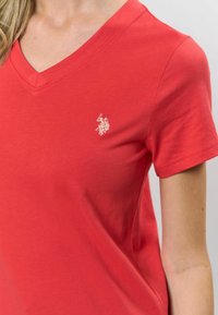 Sarkana kokvilnas V-veida kakla T-kreklu ar īsām piedurknēm un mazu izšuvumu pa kreisi uz krūtīm. Gluda tekstūra, pieguļošs fason.