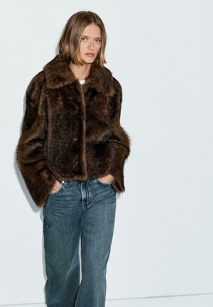 Woman wearing a brown faux fur jacket with a wide collar and blue jeans, standing with hands in pockets against a white background.