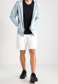 Sweat à capuche zippé bleu clair porté sur un t-shirt noir, associé à un short blanc et des baskets noires. Le sweat à capuche comprend des poches latérales et une coupe décontractée.