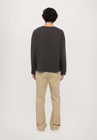 Dlhý rukáv šedohnedej košele s voľným strihom, v kombinácii so svetlobežovými nohavicami. Jednoduchý dizajn, jemná tkanina, štandardný okrúhly výstrih.