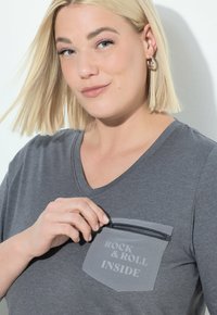 Blonde vrouw met kort haar die een borstzakje op een grijs shirt met de tekst "ROCK & ROLL INSIDE" openritst, met een lichte glimlach.