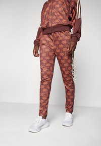 Träningsoverall i maroon med orange palmtryck, med vita sidostråk och ribbade ärmar. Matchad med vita sportskor.