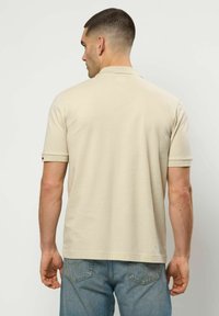 Rövid hajú férfi bézs rövid ujjú galléros pólóban és kék farmerral, hátrafele nézve, egyszínű világos háttér előtt.