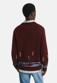 Bordeauxfarbener Strickpullover mit floralem Design am Saum, der pinke Blumen und grüne Stängel zeigt. Mit geripptem Kragen und Bündchen, über einem gestreiften Hemd getragen.