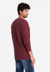 Uomo con capelli ricci che indossa una camicia a maniche lunghe color bordeaux e jeans blu, in piedi e che guarda di lato su uno sfondo bianco.