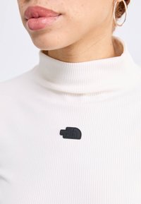 Balta stora riebalų struktūra apykaklė viršutinė dalis su mažo juodo tekstūruoto audinio žyme su "The North Face" logotipu kairėje pusėje.