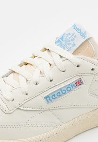 Baskets en cuir blanc avec finition texturée, présentant des accents beiges, des lacets plats et un logo Reebok bleu sur la languette et le panneau latéral.