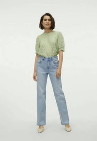 Ανοιχτό πράσινο πλεκτό τοπ με κοντό μανίκι και υφή, συνδυασμένο με ψηλόμεσα ανοιχτό μπλε τζιν και μπεζ παπούτσια με μυτερή μύτη.