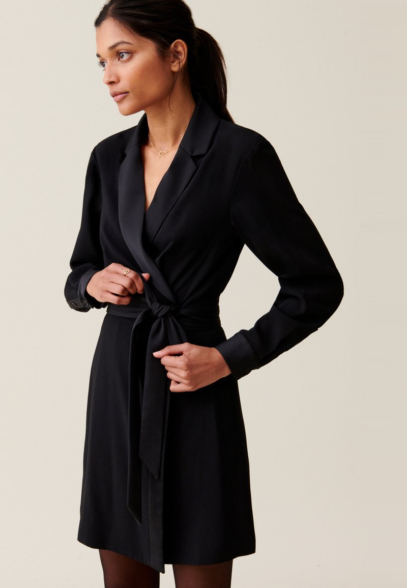 Vestito avvolgente nero realizzato in tessuto morbido, con profondo scollo a V, maniche lunghe e cintura da annodare in vita. Tessuto liscio con accenti minimi.