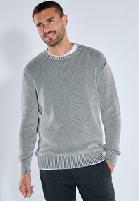 Grauer gerippter Pullover mit runder Halslinie, langen Ärmeln und gestreiften Bündchen. Über einem weißen Hemd getragen, das ein strukturiertes Strickdesign aufweist.