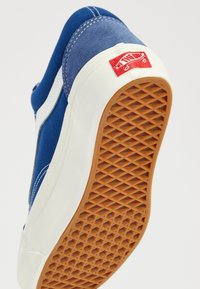 Sneaker en toile bleue avec accents blancs, capuchon de protection en caoutchouc et semelle en gomme texturée. Il présente une étiquette de logo rouge près du talon.