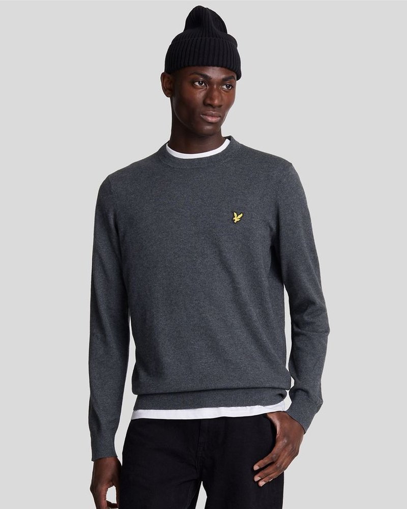 Pull en tricot gris foncé avec un col rond, des poignets et un ourlet à côtes. Présente un petit logo jaune sur la poitrine. Porté par-dessus une chemise blanche.
