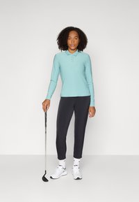 Koszulka polo z długim rękawem w jasnoniebieskim kolorze z guzikami, zestawiona z ciemnymi legginsami i białymi trampkami, trzymająca kij do golfa. Prosty i funkcjonalny design.