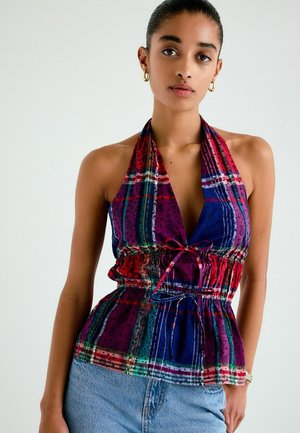 Kobieta nosząca złote kolczyki koła oraz kolorowy top na szyję z wiązaniem w talii, w połączeniu z jasnoniebieskimi dżinsami, na neutralnym tle.