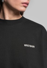 Černá bavlněná mikina s žebrovaným výstřihem a malým bílým vyšitým logem "WRSTBHVR" na hrudi.