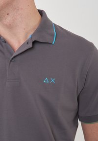 Polo grigio con colletto classico, rifiniture turchesi e logo ricamato blu sul lato sinistro del petto. Maniche corte con accenti verdi.