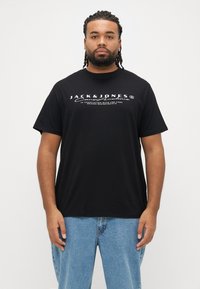 Musta puuvillainen t-paita pyöreällä kaula-aukolla, lyhyillä hihoilla, jossa valkoinen painettu logo rinnassa. Klassinen istuvuus, pehmeä tuntuma.