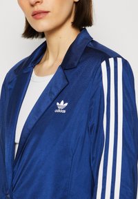 Modrá saténová bunda s designem blejzru, s bílými pruhy na rukávech a malým bílým logem Adidas na hrudi. Hladká textura.