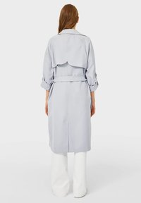 Kobieta w jasnoszarym płaszczu z paskiem i podwiniętymi rękawami, w szerokich białych spodniach, stojąca tyłem.