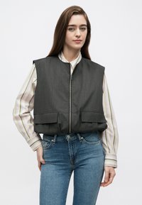 Colete cinzento acolchoado com fecho de correr à frente, grandes bolsos frontais e sem mangas, usado sobre uma camisa às riscas e jeans azuis. Textura do tecido lisa.