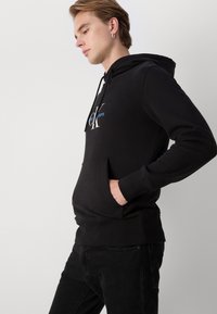 Felpa nera con cappuccio realizzata in tessuto morbido, dotata di tasca frontale, polsini a coste e design del logo in bianco e blu sul petto.