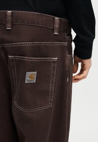 Καφέ παντελόνι από τζιν με εμφανείς λευκές ραφές και μια ετικέτα με το λογότυπο Carhartt στην πίσω τσέπη, φορεμένο με μαύρο μπλουζάκι μακρυμάνικο.