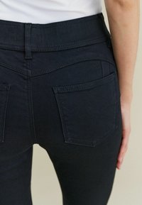 Pritisnjeni posnetek osebe v črnih prilegajočih se hlačah, ki prikazujejo zadnja žepa in zanke za pas, z белo majico, vtaknjeno v hlače.