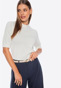 Witte, kortemouw gebreide top met een hoge hals, gecombineerd met marineblauwe pantalon. Een beige riem met een metalen gesp accentueert de outfit.