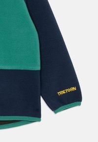 Fleecejakke med mørkeblå og grønne paneler, med "TRETORN" brodert i gult på ermet. Glatt tekstur, atletisk design.