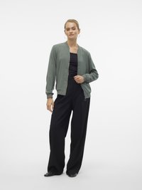 Femme aux cheveux attachés portant des boucles d'oreilles en anneau, une veste bomber vert clair, un haut noir, un pantalon large noir et des chaussures plates noires.