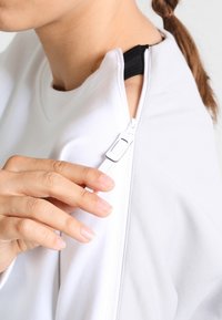 Sudadera blanca con un detalle de cremallera en el hombro. La textura parece suave, con un acento negro cerca de la cremallera. Mano mostrando un agarre sobre la cremallera.