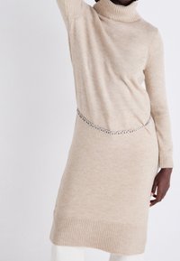 Femme portant une robe beige en tricot à col roulé, à manches longues et avec une ceinture en chaîne argentée, sur fond blanc uni.