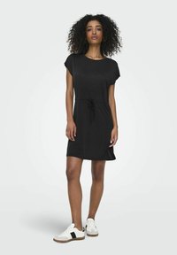 Robe noire à manches courtes avec taille à cordon, coupe décontractée et texture lisse. Portée avec des baskets blanches à accents noirs.