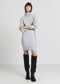 Abito maglione grigio lavorato a maglia con collo alto, caratterizzato da un motivo a trecce e lunghezza a metà coscia, abbinato a stivali neri al ginocchio.