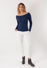 Maglione blu navy a maniche lunghe con design off-shoulder, abbinato a pantaloni bianchi slim-fit e stivaletti neri alla caviglia con dettagli in zip.